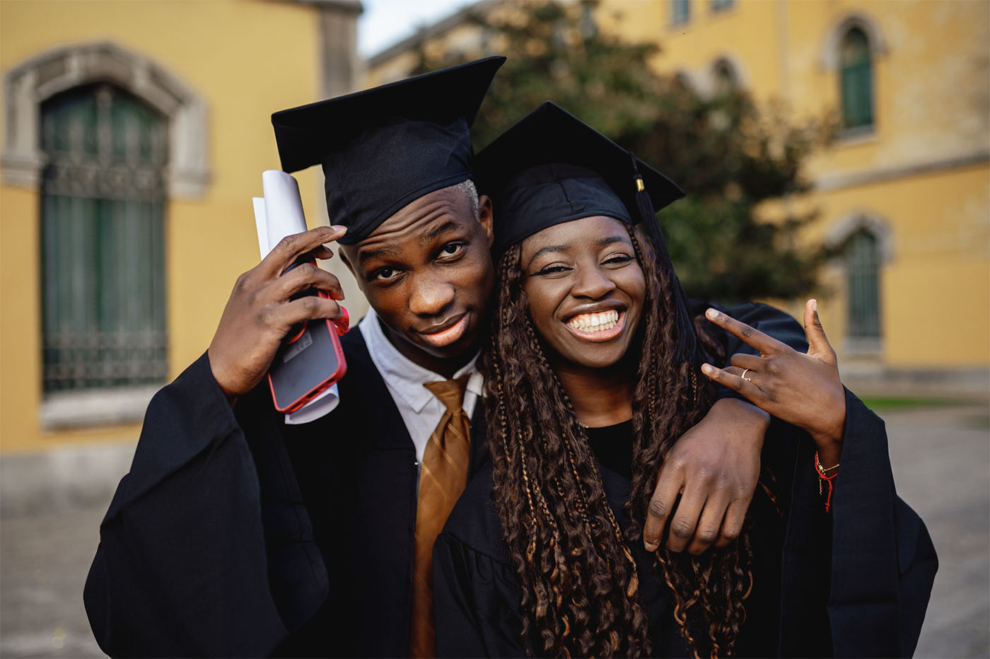 Two young black students wearing their graduation gowns are hugging and smiling at the university campus on graduation day